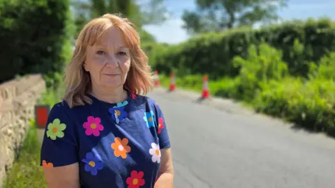 Paul Moseley/BBC Wendy Allsop is standing to the side of a country road. She is wearing a blue top, patterned with different coloured flowers.