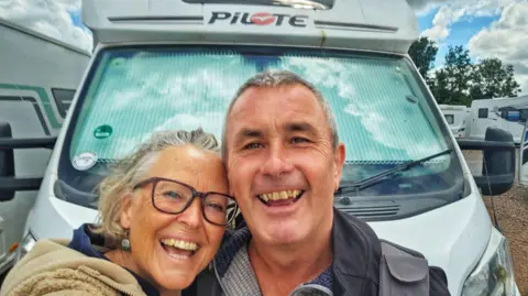 Image of a tanned couple who are standing in front of a motorhome and smiling at the camera. The woman is wearing glasses and has silver earrings and a beige hooded top.  The man has grey short hair and is wearing a black jacket.  In the background can be seen a number of motorhomes.