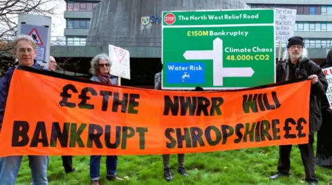 Three people standing on grass, holding up a large orange banner which reads 'the NWRR will bankrupt Shropshire'.
