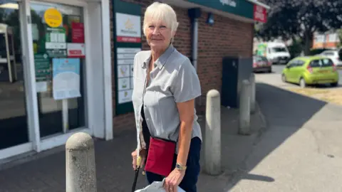 An elderly woman with white blonde hair and a walking stick stands in front of the local shop - a single story red brick building with the Morrisons log in the background.