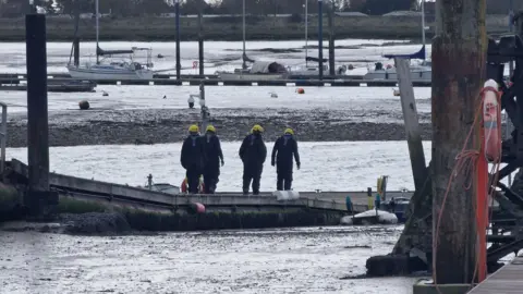Essex Police Four people with their backs to the camera wearing dark outfits and yellow helmets. They are overlooking what appears to be a backwaters. Boats are in the background and wooden structures are in the foreground. 