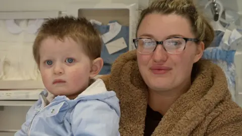 Jamie Niblock/BBC A woman in a fluffy brown hooded top is stood inside a business with baby clothes on the shelves behind her. She is holding a baby in a blue outfit.