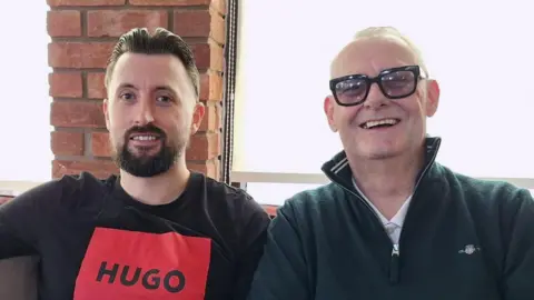 Father and son Mark (right) and Aaron Bond. Aaron has brown hair and beard is wearing a black t-shirt with a red square with the 'Hugo' written inside it in black. Mark has white receding hair and black glasses and wears a white shirt with a dark green zipped up jumper. They are sitting inside a building in front of windows with cream blinds and a brick wall.