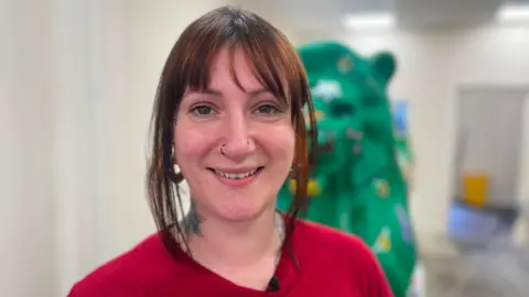 Amy is smiling at the camera. She has brown hair which is tied up. She has a fringe and pieces of hair framing her face. She has a red top on. Behind her, is a green lion she has painted which is inspired by wildlife.