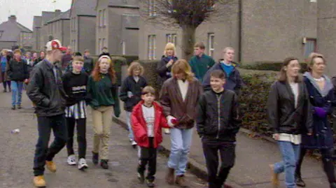 A photo of a protest in Raploch in the 1990s. Lots of local people are walking down the street with houses in the background. 
