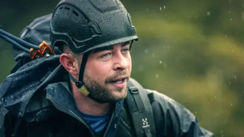 A man with stubble and blue eyes wears a dark hard hat, backpack and jacket. There's a blurred, green background and there are bursts of rain.
