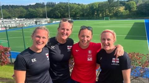 Lucy Field and three of her teammates smile at the camera in front of a hockey pitch. They have their arms around each other and three of them are wearing blue T-shirts and one is wearing a red T-shirt. 