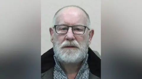 Northumbria Police Mugshot of Wilson. He has a thick white beard and bald head and is wearing black-framed glasses