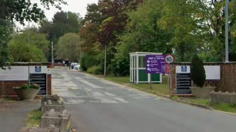 A road runs between two walls, with a number of signs on both walls showing that it is a prison. A larger sign behind the wall on the right says HMP and YOI Sudbury