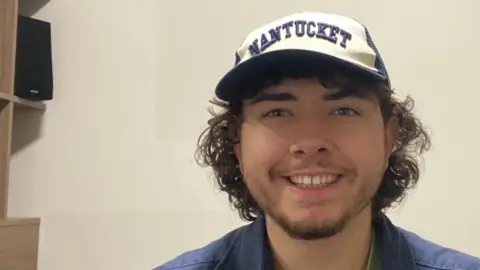 Jordan Andrews A young man wearing a cap, smiling, sitting on a bed in student accommodation