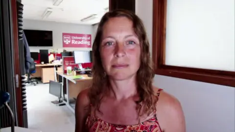 Professor Hannah Cloke stood in the University of Reading office wearing a summery top and looking at the camera. Behind her is desks with computers resting on them and a University of Reading sign on the wall.