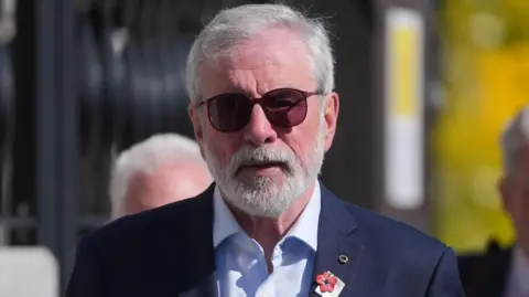 PA Media Gerry Adams with short grey hair and facial hair. He is wearing a navy suit with a light blue shirt collared shirt. He has round, sunglasses on and there are badges on his suit jacket. Behind him are black gates.