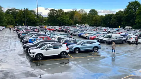 A very busy open-aired car park, with people, pay machines and street lighting visible. Trees outline the car park and rain has fallen.
