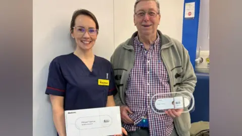 Rachel Hilmo wearing a dark blue clinical outfit holds a white cardboard box while standing with a patient, who is an older man wearing a green cardigan and a checked red and blue shirt. He is holding the wearable patch which is oval in shape and transparent on the edges and white in the centre.