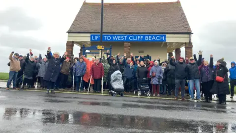 Carla Fowler/BBC A group pf protesters dressed in coast and waterproofs cheering outside a small building alongside a road. The building has a sign on it saying, lift to West Cliff beach.