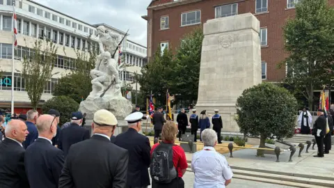 A wider view of the service at the monument. The Boer war memorial stands in front of the Cenotaph and depicts two uniformed soldiers in a field action stance.