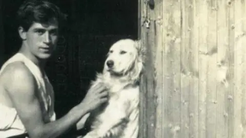 Dorset Museum Black and white photo of David Brynley as a young man fussing a light-coated retriever-type dog. He has dark hair with a long fringe and is wearing a sleeveless vest.