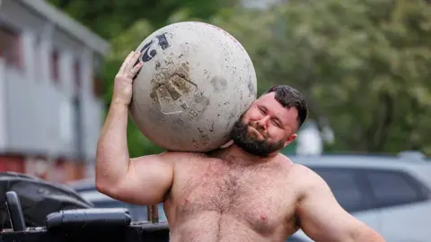 Corporal Rhys Calland lifts a huge spherical stone on his shoulder, with a grimace on his face. He is topless and cars and trees can be seen in the background. 