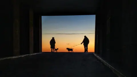 Bruce Taylor The image shows two people, a woman and a man, walking their dogs along a paved path. The path leads out of a shadowy, arched passageway into a bright, golden sunset. Both figures are in silhouette against the setting sun. Behind them, the sky is a mix of orange, pink and blue.