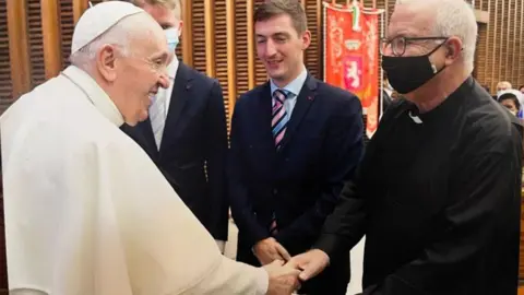 A canon on the right wearing a mask, shaking hands with the Pope.