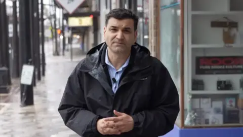 BBC News correspondent Gordon Corera wears a raincoat and looks into the camera