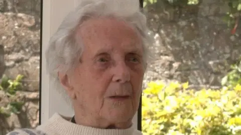 BBC Valerie Guy looking past the camera seated in front of a window that frames a stone wall and bush outside