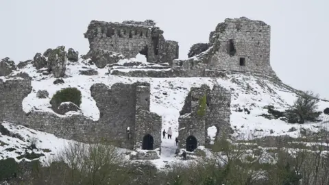 PA A ruined castle on a hilltop which is covered by snow. Walkers who are wrapped up warmly are walking among the ruins through the snow.