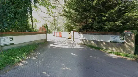 Google The entrance to the site of Ailwyn Hall, which are two brick walls either side of a brick driveway, lined with trees. You can't see the actual care home. There are signs on each wall saying Ailwyn Hall Care Home, Ashley Care Group.