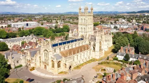 Drone shot of Gloucester Cathedral in the sunshine. Images shows nearby houses and businesses and green spaces.