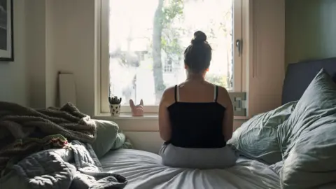 A generic picture of a girl sitting on a bed and looking out of a window. She has her back to the camera.