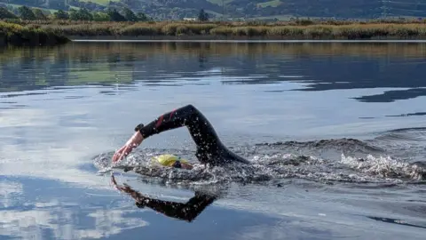 Piotr Gudan Image shows Calum swimming in the River Tay between Perth and Newburgh in September this year. The water is ferry still. He is submerged with just one side of his head and one arm and shoulder reaching out of the water. He is wearing a yellow swim cap, black goggles and a dark wetsuit.