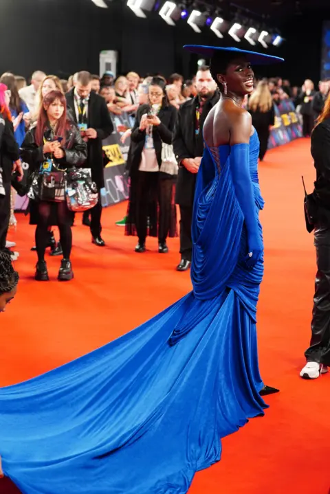 PA Media Jodie Turner-Smith on the red carpet in a striking blue gown with a long train and a matching circular flat hat