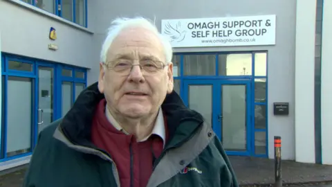 Michael Gallagher. He has white hair, wearing glasses, a dark rain coat with purple jacket, purple jumper and white shirt. He is standing in front of a building with a sign for 'Omagh Support & Self Help Group' above the doorway.