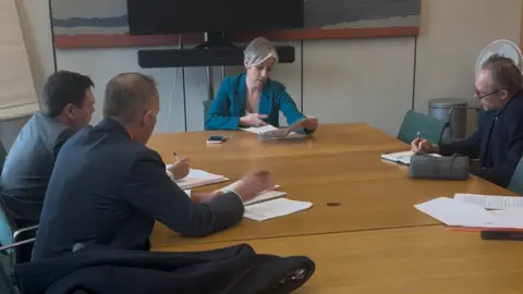 Daisy Cooper Daisy Cooper sitting at the end of a table, meeting a number of men in suits.