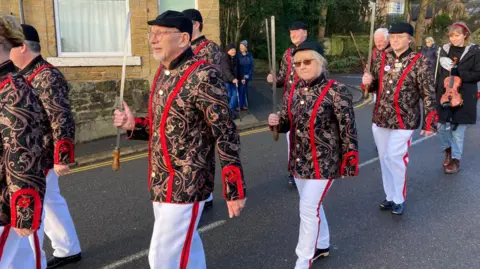 Eight sword dancers parade through a street. They are wearing their paisley jackets, black caps and white trousers, which have a red stripe. A woman with a fiddle follows them. Spectators watch from the pavement.