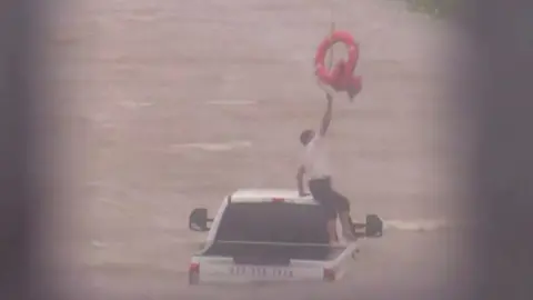 A man sits on a half-submerged truck and reaches for a life ring above his head