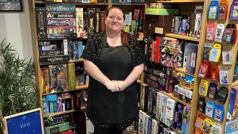 Lindsey Bird wearing a black top and a black apron standing in front of a shelf full of games