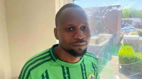 Mutasim Mohammed is a black man with short black hair and beard. He is standing in front of a smashed window on his home. He is wearing a Northern Ireland football top