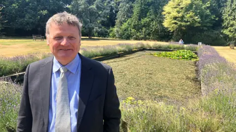 Richard Edwards/BBC A man stands in front of an ornamental pond, set within a large lawn ringed by trees. He is wearing a suit and tie, has short brown hair and is smiling towards the camera.