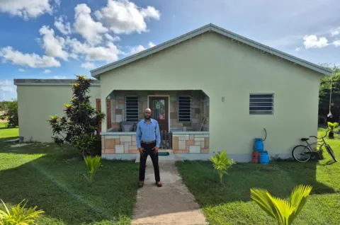 Courtesy Dwight Benjamin Dwight Benjamin, wearing a blue shirt and black jeans, standing in front his his home. Behind him the porch and entrance door can be seen. A bicycle is leaning against the front wall of the. Some gas cylinders are stacked in front of the house. To the left of the house, the newly built extension which serves as a shelter can be seen.