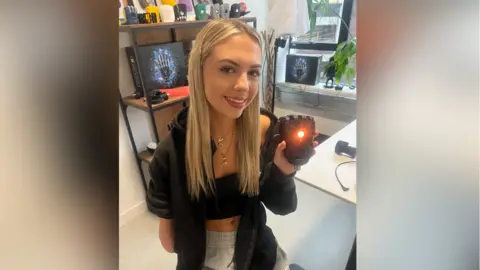 Sadie Sadie, who has long blonde hair and is wearing grey tracksuit bottoms, a black top and a black jumper, is holding up the prosthetic hand, which has an orange light flashing from the centre of it. 
