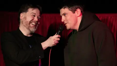 Brendan O'Connor Two men backed by a silk red curtain behind them. One on the left has a greying beard and short dark hair dressed in all black. He holds up a microphone with a red cable to the man on the right, who has short brown hair. He is wearing a dark green hoodie.