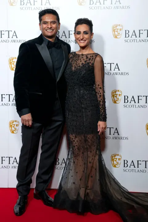 PA Media Dr Sandesh Gulhane, in a black suit, and Dr Punam Krishan, in a long black dress with lace train, stand in front of a Bafta Scotland backdrop