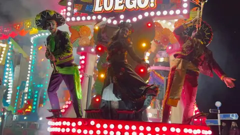 Three performers on a carnival float. They are all wearing face paint to mimic Day of the Dead skulls. 