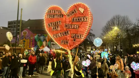 A crowd of people, in the foreground a woman is carrying a giant red illuminated heart with the words "Bristol together, home, Banksy, street art, music" and a few other phrases on it that describe the city