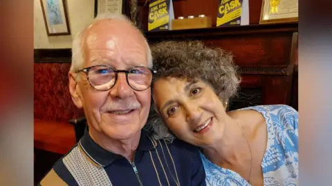 A man and a woman advancing in years, the man has white hair and moustache and glasses, the woman has dark, greying curly hair. They are hugging and smiling at the camera.