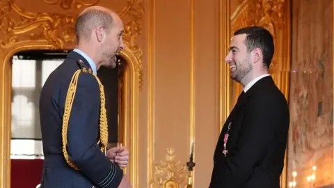 PA Media Luke Humphries on the right of the image wearing an all black suit and smiling as he talks to Prince William on the left who is dressed in military attire. They are stood in a grand room in Windsor Castle with a host of people in the background.