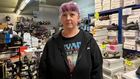 Simon Thake/BBC A woman in a black hoodie and purple hair stands in front of displays of shoes.