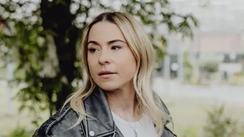 LDRS A woman with long blonde hair wearing a white T shirt and leather jacket turns to face away from the camera