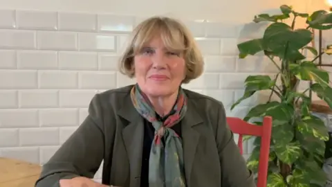 Denise McKay with blonde hair, wearing an olive blazer and a pashmina. She is sitting at a wooden table on a red chair looking at the camera.